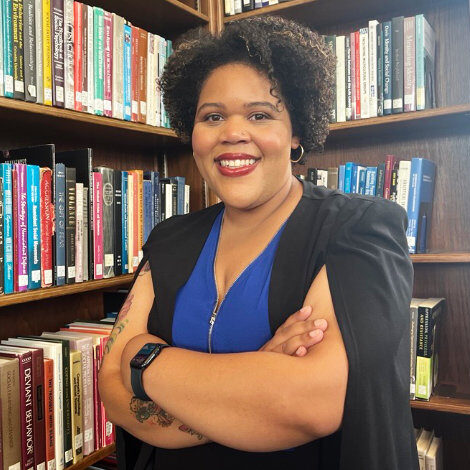 Portrait of a smiling woman with short curly hair, wearing a blue top and black vest, arms crossed in a library aisle.