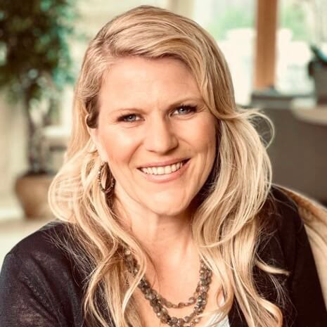 Portrait of a smiling blonde woman wearing a dark cardigan and layered necklace, indoors with natural light on her face.